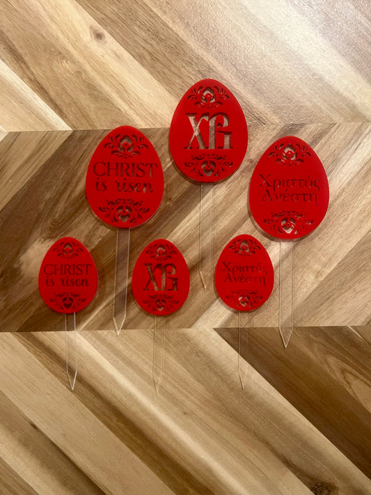 A red Easter egg-shaped decoration to put on top of kulich or other Pascha or Easter dish, with the Russian Slavonic XB, English text Christ is Risen or Greek text Christos Anesti and floral cutouts. the design is cut out on red acrylic which is attached to a clear acrylic stake available in two sizes.