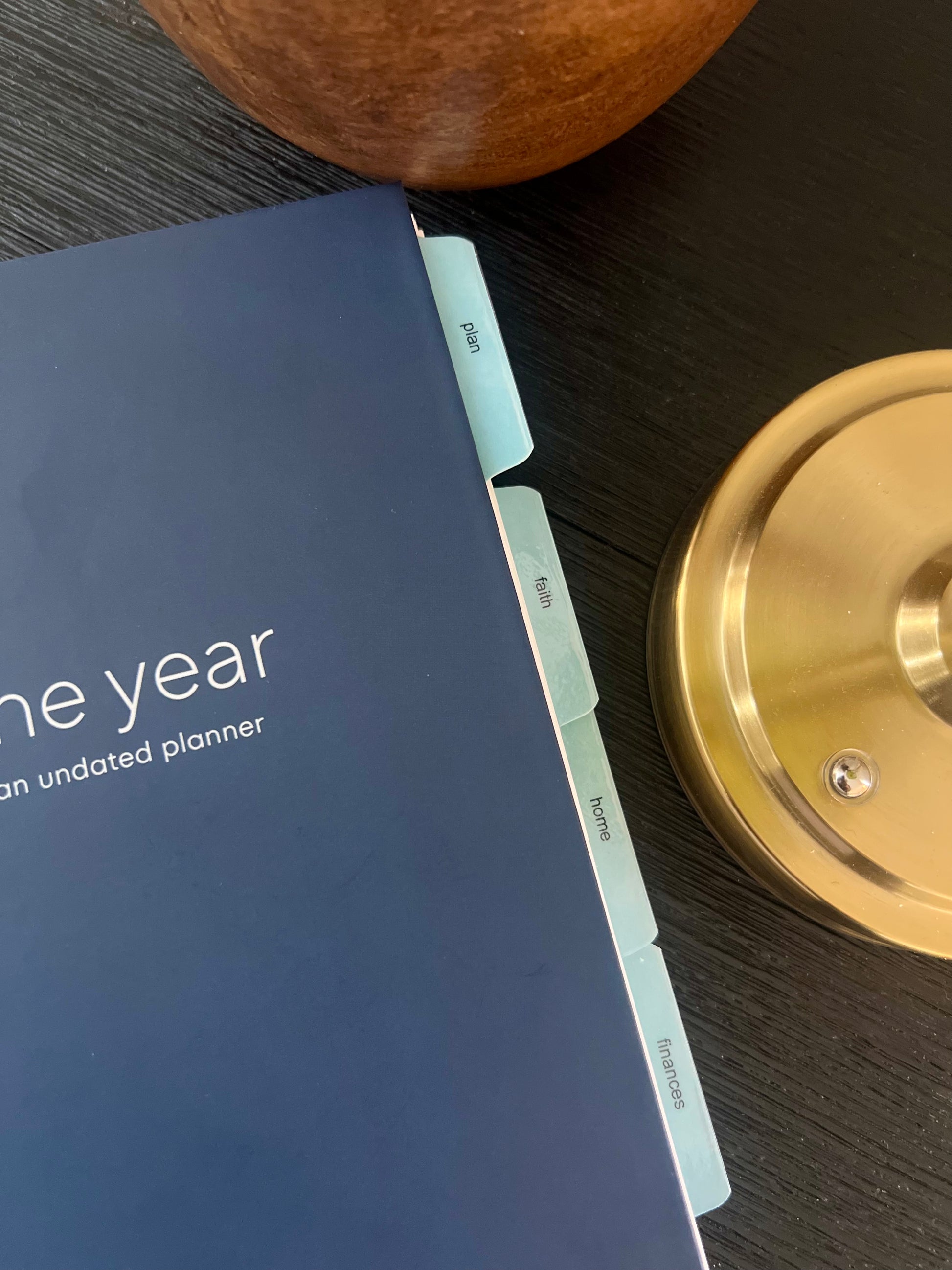 A close-up of a navy blue one-year undated Orthodox life planner with light blue tabs on a dark wood surface, alongside a wooden bowl and a brass candlestick base.