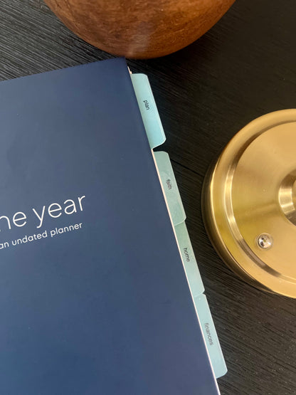 A close-up of a navy blue one-year undated Orthodox life planner with light blue tabs on a dark wood surface, alongside a wooden bowl and a brass candlestick base.