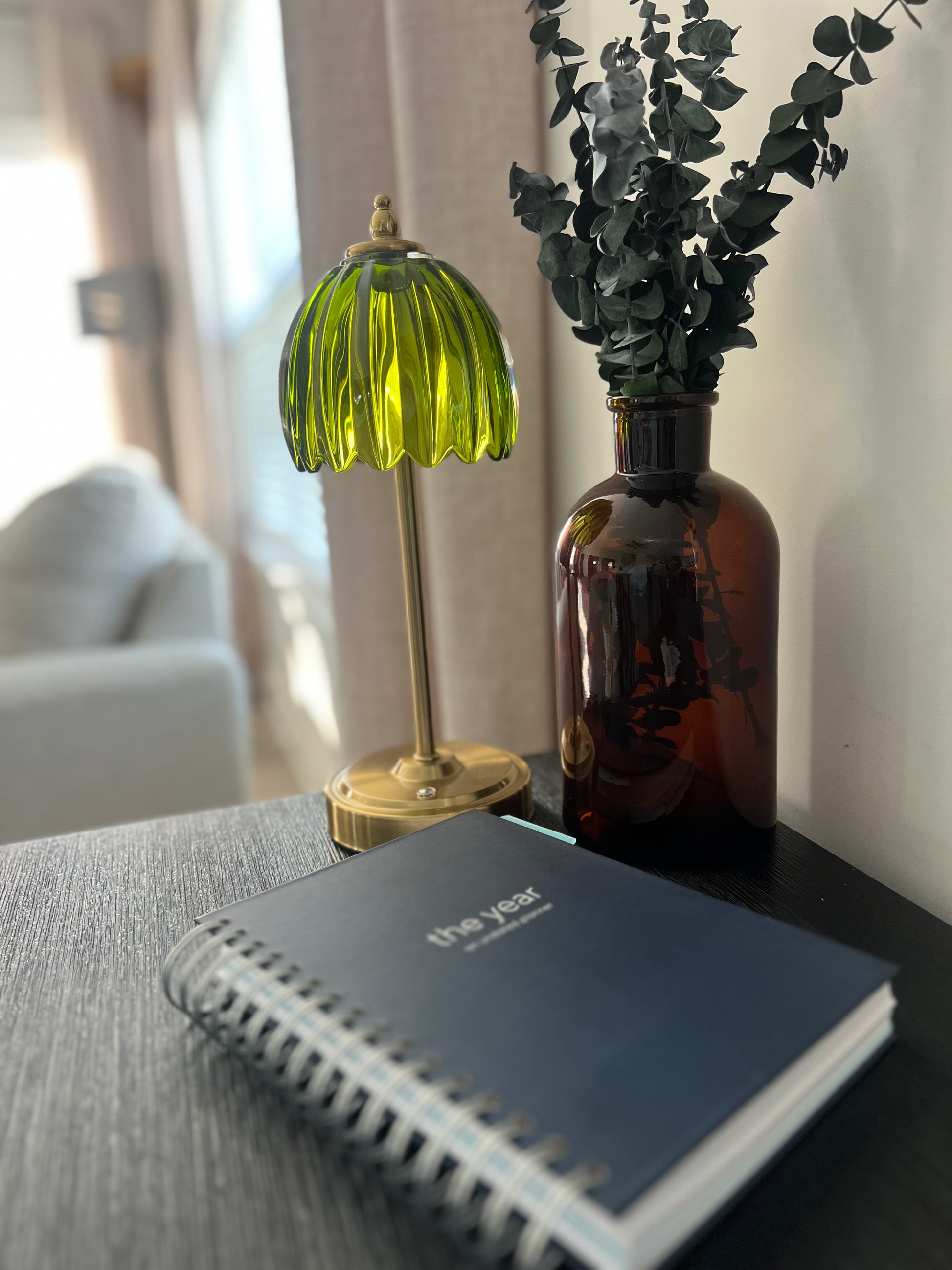 A navy blue spiral Orthodox undated planner with silver wire binding in the foreground, with a small green ribbed glass table lamp and an amber glass vase with dried greenery behind it on a dark tabletop.